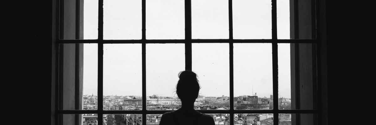 A black and white shot of a lonely female standing in front of the windows looking at the buildings